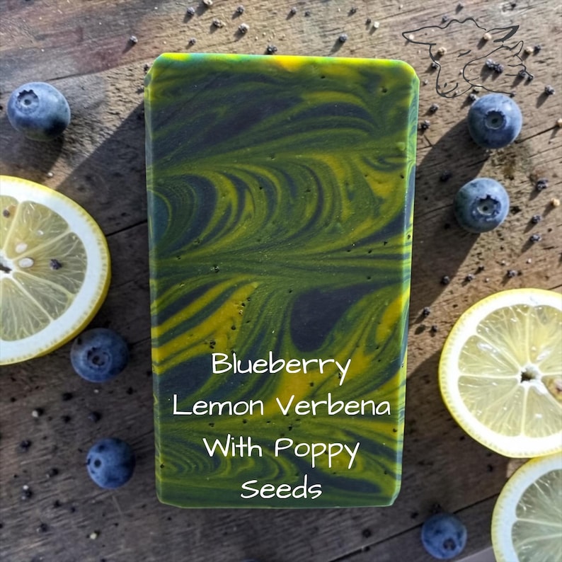 Puede incluir: Una pastilla de jab&oacute;n rectangular con un patr&oacute;n jaspeado de carb&oacute;n oscuro y amarillo vibrante. El texto en el jab&oacute;n dice: Blueberry Lemon Verbena With Poppy Seeds. La pastilla est&aacute; rodeada de ar&aacute;ndanos frescos y rodajas de lim&oacute;n amarillo.
