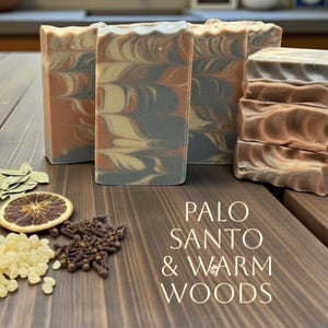 May include: Artisan soap bars with a swirled design in shades of gray, beige, and rust. The bars are arranged on a wooden surface with dried botanicals and the text "PALO SANTO & WARM WOODS".