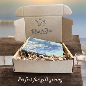 May include: A rectangular bar of soap with a marbled blue, grey, and cream pattern nestled in a brown cardboard box filled with shredded paper. The inside lid features a printed logo with a goat illustration and the text Tallow & Shea Goat Milk Soap.