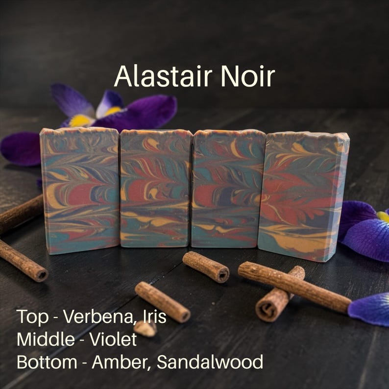 May include: Four bars of soap with a colorful, swirled design in shades of blue, red, and gold. The soap is labeled "Alistair Noir" with scent notes of Verbena, Iris, Violet, Amber, and Sandalwood. Cinnamon sticks and purple flowers are scattered around the soap.