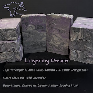 May include: Several bars of handmade soap with a swirling design in shades of gray, purple, and white are stacked and arranged on a dark surface. The text "Lingering Desire" is visible, along with scent notes: Norwegian Cloudberries, Coastal Air, and Blood Orange Zest.