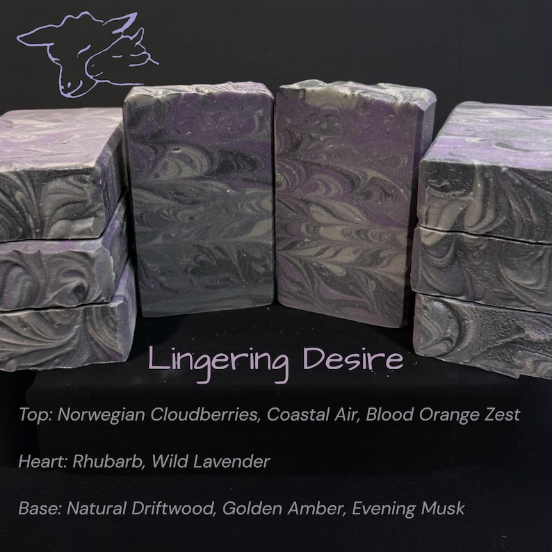 May include: Several bars of handmade soap with a swirling design in shades of gray, purple, and white are stacked and arranged on a dark surface. The text "Lingering Desire" is visible, along with scent notes: Norwegian Cloudberries, Coastal Air, and Blood Orange Zest.