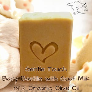 May include: A rectangular bar of light tan soap featuring an embossed heart in the center. The text on the soap reads: Gentle Touch, Baby Bastille with Goat Milk, 80% Organic Olive Oil. A line drawing of two sheep heads is visible above.