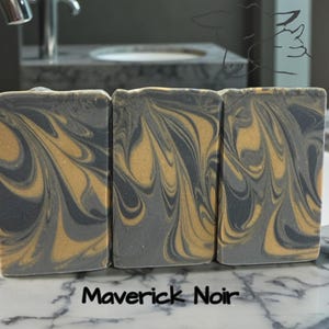 Puede incluir: Tres barras de jab&oacute;n rectangulares con un patr&oacute;n jaspeado de remolinos gris carb&oacute;n oscuro y oro met&aacute;lico. El texto Maverick Noir est&aacute; impreso en negro en la parte inferior. Un tenue dibujo lineal de una cabeza de rinoceronte es visible arriba a la derecha.