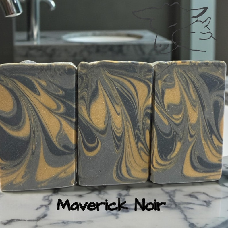 Puede incluir: Tres barras de jab&oacute;n rectangulares con un patr&oacute;n jaspeado de remolinos gris carb&oacute;n oscuro y oro met&aacute;lico. El texto Maverick Noir est&aacute; impreso en negro en la parte inferior. Un tenue dibujo lineal de una cabeza de rinoceronte es visible arriba a la derecha.