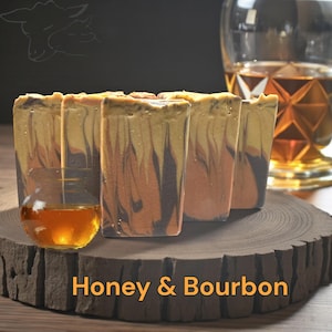 May include: Five rectangular bars of soap with a swirled pattern of brown, yellow, and orange hues, arranged on a wooden slab. The text "Honey & Bourbon" is displayed in orange. A glass of liquid is in the foreground.