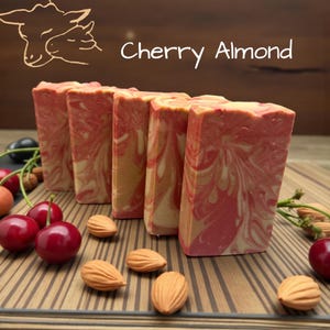 Puede incluir: Cinco barras rectangulares de jab&oacute;n con un patr&oacute;n de remolinos jaspeados en rosa, rojo y crema. El texto Cherry Almond est&aacute; impreso sobre el jab&oacute;n, junto a una ilustraci&oacute;n lineal de dos ovejas. Cerezas rojas frescas y almendras est&aacute;n esparcidas.