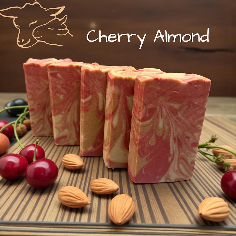 Puede incluir: Cinco barras rectangulares de jab&oacute;n con un patr&oacute;n de remolinos jaspeados en rosa, rojo y crema. El texto Cherry Almond est&aacute; impreso sobre el jab&oacute;n, junto a una ilustraci&oacute;n lineal de dos ovejas. Cerezas rojas frescas y almendras est&aacute;n esparcidas.