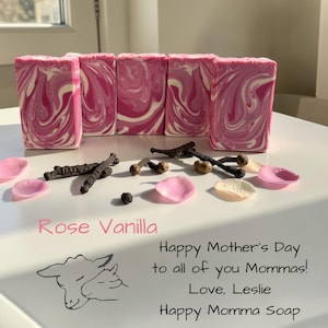 May include: Pink and white swirled soap bars with rose petals and vanilla beans. The text "Rose Vanilla" is displayed, along with a Mother's Day message and a sheep illustration. The soap is from Happy Momma Soap.