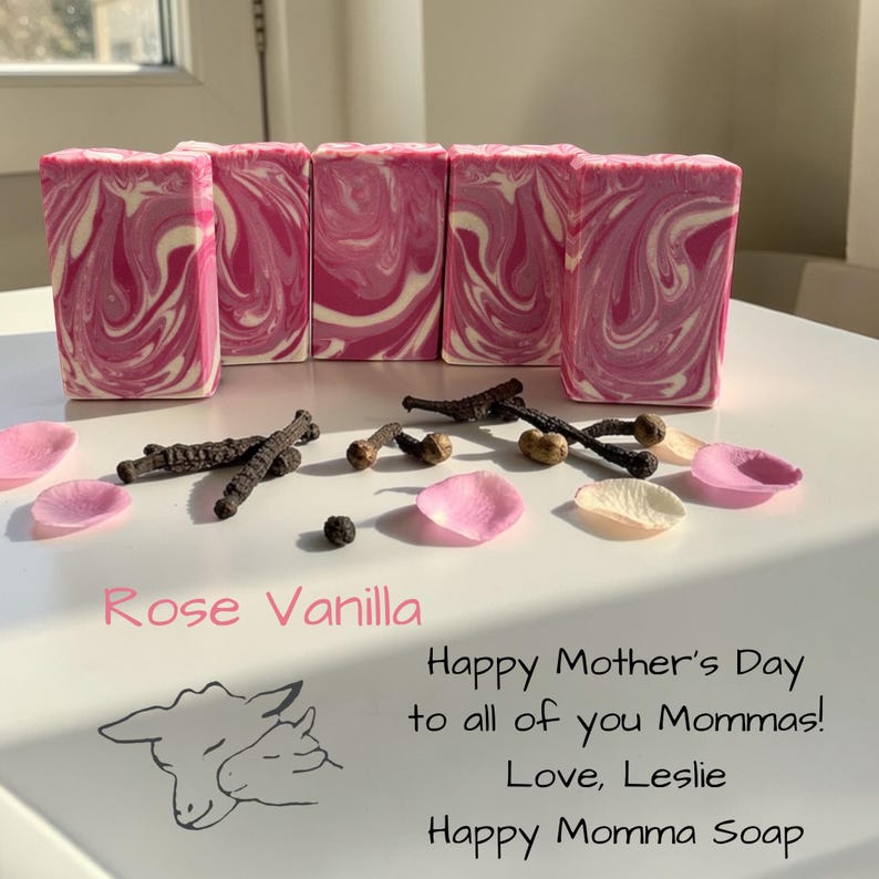 May include: Pink and white swirled soap bars with rose petals and vanilla beans. The text "Rose Vanilla" is displayed, along with a Mother's Day message and a sheep illustration. The soap is from Happy Momma Soap.