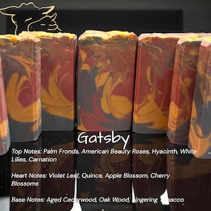 May include: Artisan soap bars with a marbled design in shades of red, gold, and black. The word "Gatsby" is displayed in white script. The soap's top, heart, and base notes are listed below the name.