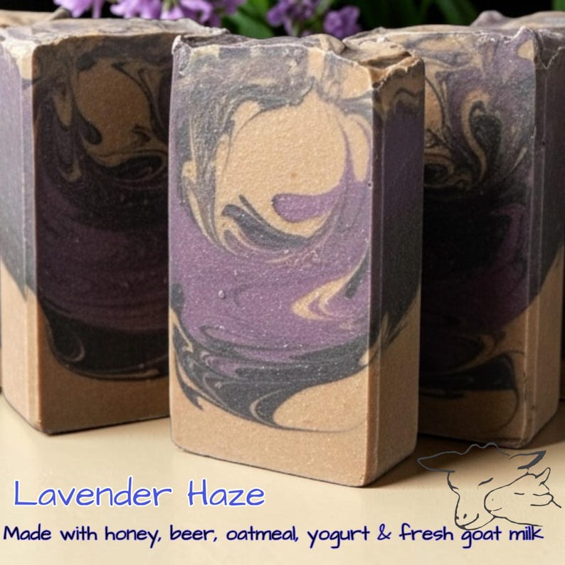 May include: Three rectangular bars of soap featuring a marbled pattern of deep purple, black, and tan colors. The text Lavender Haze is printed in blue, followed by the text Made with honey, beer, oatmeal, yogurt & fresh goat milk.