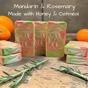 May include: Rectangular bars of soap with a marbled swirl pattern in shades of orange, green, and white. The text Mandarin & Rosemary Made with Honey & Oatmeal is visible above the soap. Fresh rosemary sprigs and whole oranges are present.