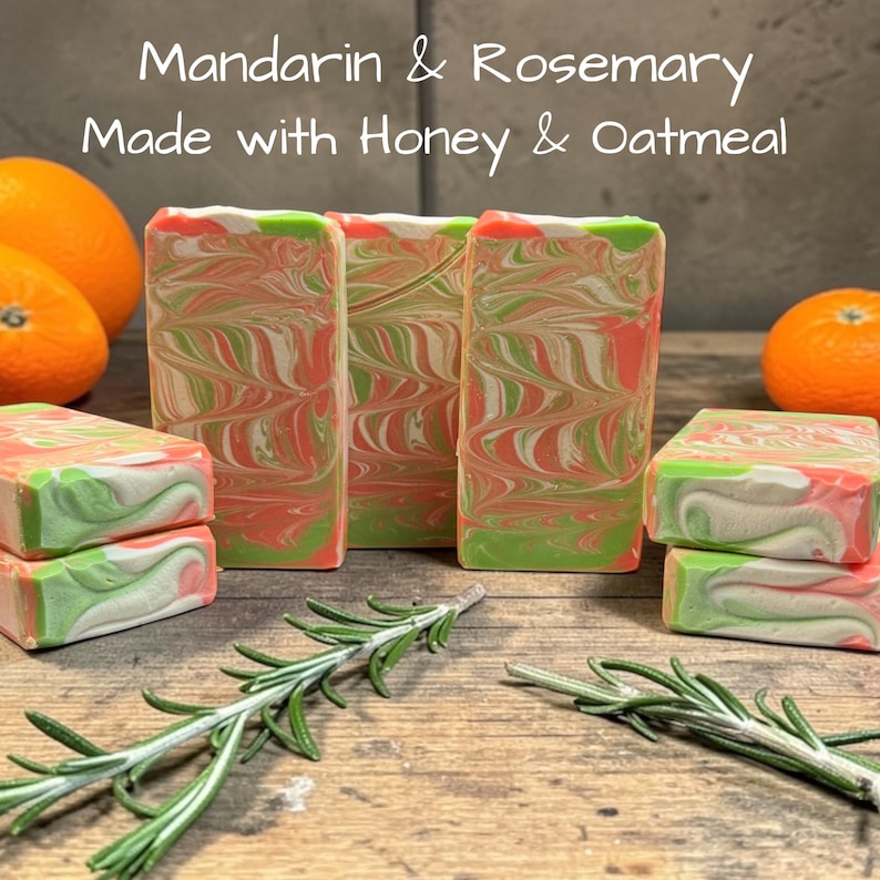 May include: Rectangular bars of soap with a marbled swirl pattern in shades of orange, green, and white. The text Mandarin & Rosemary Made with Honey & Oatmeal is visible above the soap. Fresh rosemary sprigs and whole oranges are present.