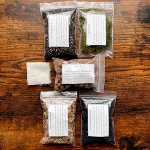 May include: A terrarium kit with soil, moss, charcoal, and other components in clear plastic bags. The kit includes Terrarium Soil, Live Moss, LECA, Sphagnum Moss, Activated Charcoal, and a cheesecloth. The items are arranged on a wooden surface.