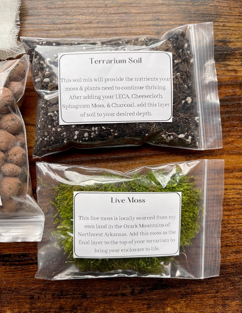May include: Two clear plastic bags containing terrarium supplies. One bag is filled with dark soil labeled "Terrarium Soil." The other bag contains green "Live Moss." Both bags have white labels with descriptive text.