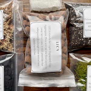 May include: A collection of terrarium supplies in clear plastic bags, including LECA (Light Expanded Clay Aggregate), moss, and other materials. The LECA bag has a label explaining its use for drainage. The items are arranged on a wooden surface.