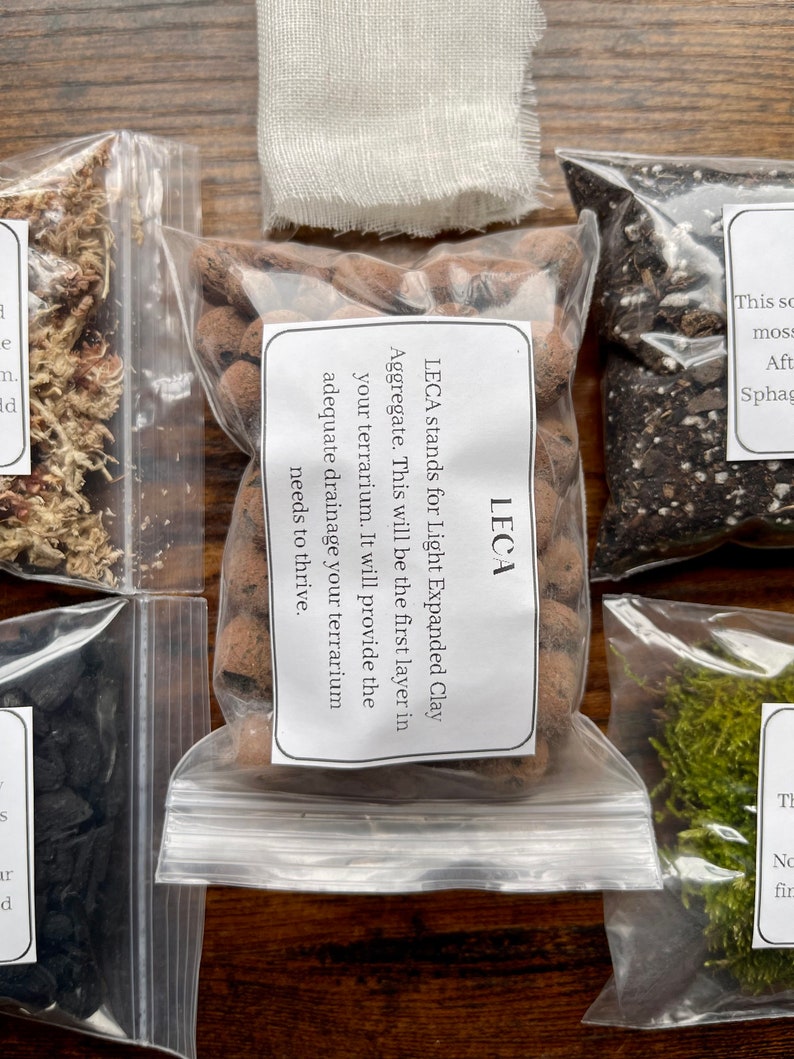 May include: A collection of terrarium supplies in clear plastic bags, including LECA (Light Expanded Clay Aggregate), moss, and other materials. The LECA bag has a label explaining its use for drainage. The items are arranged on a wooden surface.