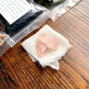 May include: Three light pink, raw rose quartz crystals rest on a small, frayed, off-white cloth. Behind the cloth are clear plastic bags containing activated charcoal and moss, with handwritten labels. The items are arranged on a wooden surface.