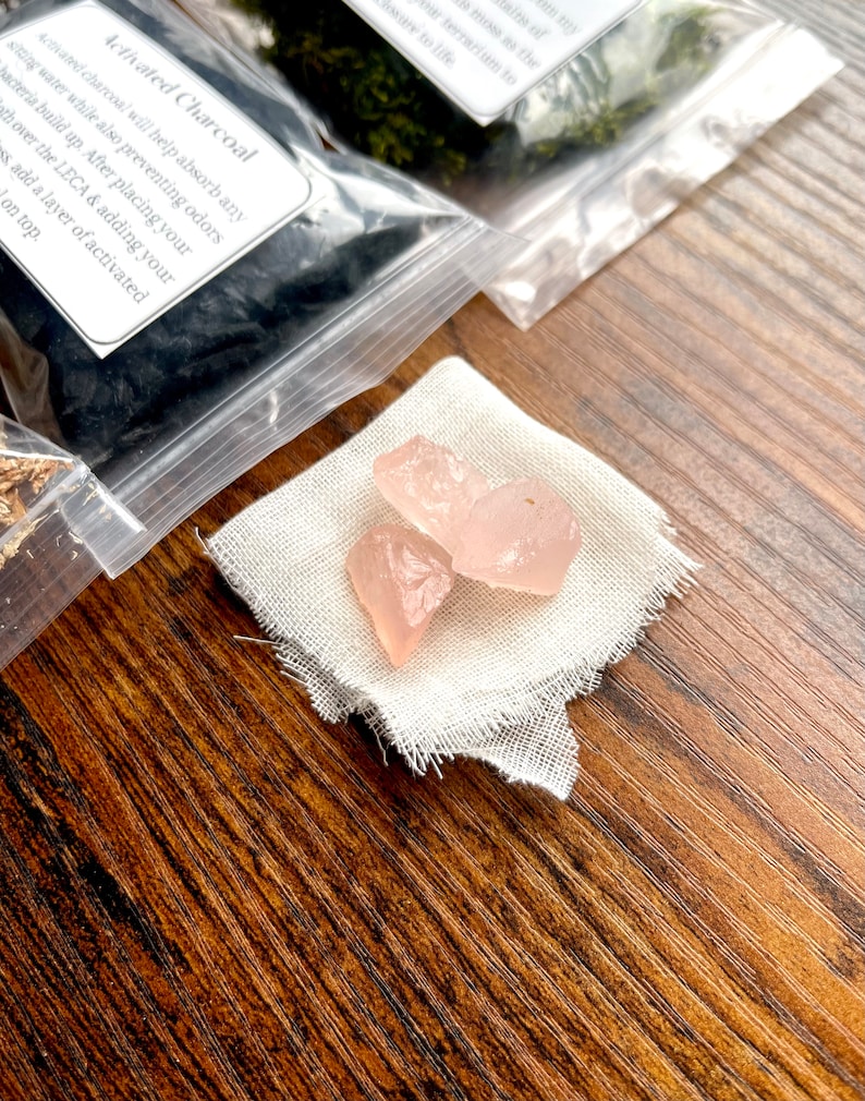 May include: Three light pink, raw rose quartz crystals rest on a small, frayed, off-white cloth. Behind the cloth are clear plastic bags containing activated charcoal and moss, with handwritten labels. The items are arranged on a wooden surface.