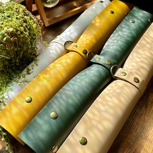May include: A collection of colorful leather pen cases in shades of yellow, green, and beige. Each case is secured with a brass snap and a leather strap with a metal ring. The cases are arranged on a wooden surface.