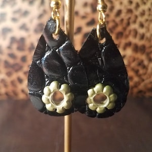 May include: A pair of black leather teardrop earrings with gold accents and a flower design. The earrings have a textured surface and a gold hook.