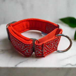May include: A bright orange dog collar featuring a silver metallic swirl pattern on the outer fabric. The collar includes a soft orange fleece lining, a sturdy metal D-ring for leash attachment, and a metal slide adjuster for sizing adjustments.