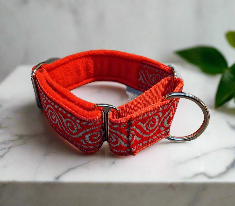 May include: A bright orange dog collar featuring a silver metallic swirl pattern on the outer fabric. The collar includes a soft orange fleece lining, a sturdy metal D-ring for leash attachment, and a metal slide adjuster for sizing adjustments.