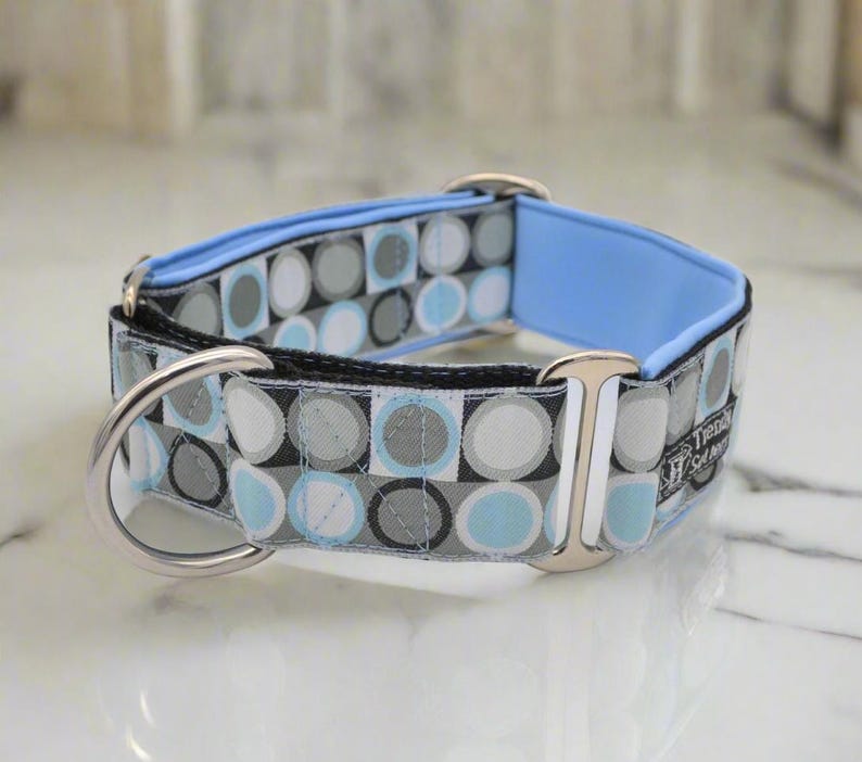 May include: A dog collar featuring a repeating geometric pattern of circles in shades of light blue, gray, white, and black. The collar includes a light blue inner lining, silver-toned metal hardware, a D-ring, and a small tag labeled Trendy Set Jerry.