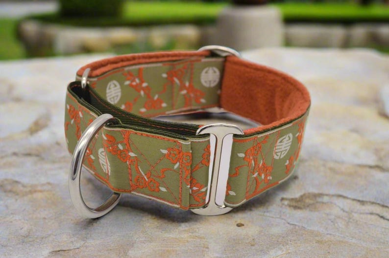 May include: A wide pet collar featuring an olive green fabric with an orange floral pattern and white circular symbols. The collar includes polished silver metal hardware, including a D-ring and a rectangular slide, with a soft orange inner lining.