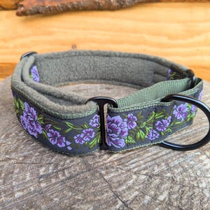 May include: A pet collar featuring a dark gray outer band with a purple and green floral pattern. The collar includes a soft olive green fleece lining, black metal hardware, and a sturdy D-ring for attachment. The design is adjustable and durable.