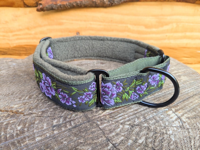 May include: A pet collar featuring a dark gray outer band with a purple and green floral pattern. The collar includes a soft olive green fleece lining, black metal hardware, and a sturdy D-ring for attachment. The design is adjustable and durable.
