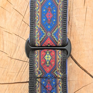 May include: A dog collar featuring a woven fabric strap with a repeating geometric pattern in red, blue, and yellow. The collar includes black plastic hardware, including a buckle and a D-ring, with black nylon webbing edges along the length.