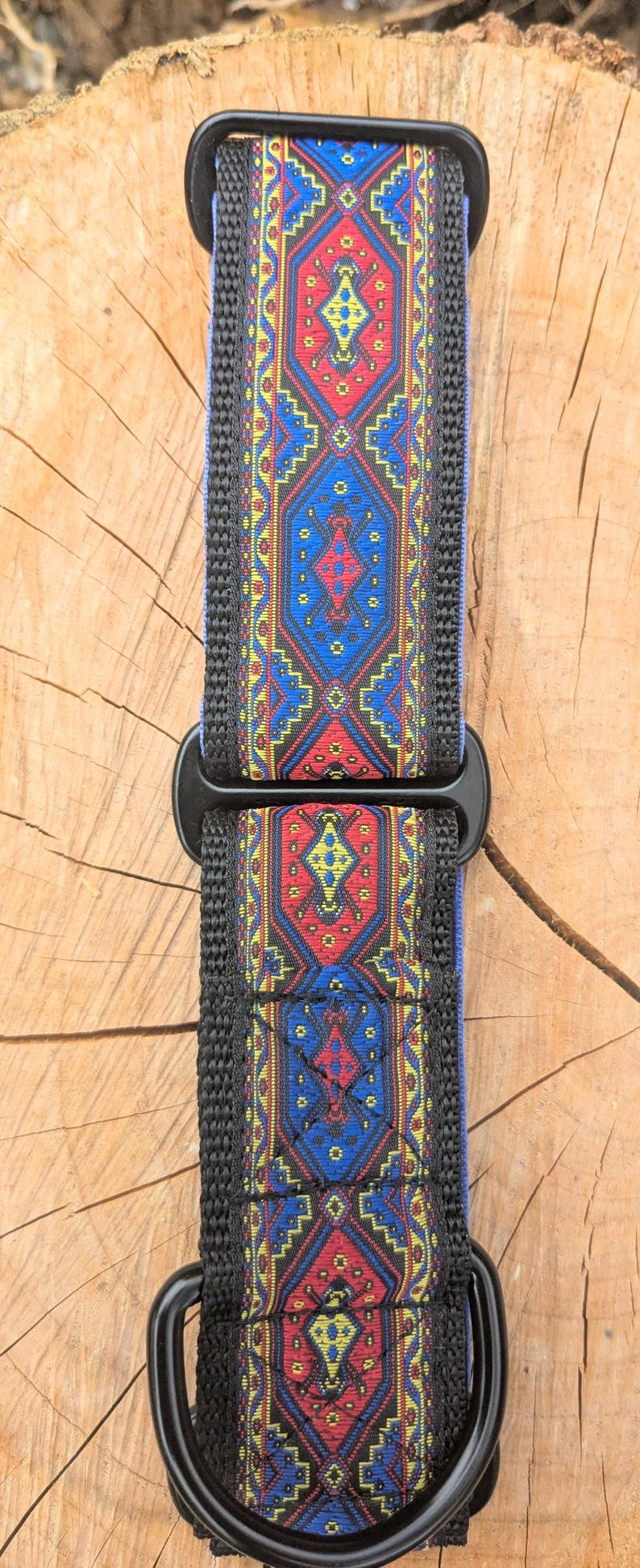 May include: A dog collar featuring a woven fabric strap with a repeating geometric pattern in red, blue, and yellow. The collar includes black plastic hardware, including a buckle and a D-ring, with black nylon webbing edges along the length.