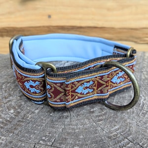 May include: A pet collar featuring a decorative woven ribbon with a dark red and gold intricate pattern, accented by light blue floral motifs. The collar has a soft light blue padded interior, black webbing, and antique brass metal hardware.