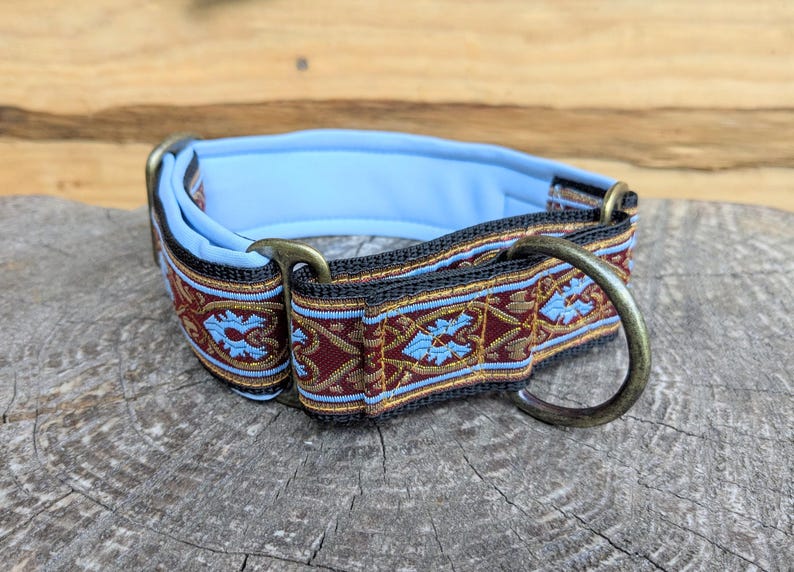 May include: A pet collar featuring a decorative woven ribbon with a dark red and gold intricate pattern, accented by light blue floral motifs. The collar has a soft light blue padded interior, black webbing, and antique brass metal hardware.