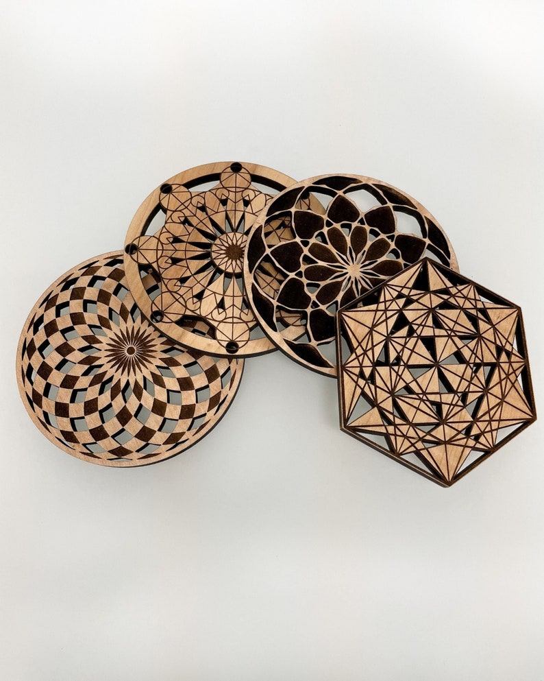 May include: Four wooden coasters with intricate geometric designs. The coasters feature various patterns, including a spiral, a floral design, and a geometric shape. The wood has a natural, light brown color, and the designs are cut out.