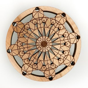 May include: A round, wooden coaster with a geometric design. The coaster features a central starburst pattern surrounded by interconnected circles and lines, all laser-cut into the light-colored wood. The design is intricate and symmetrical.