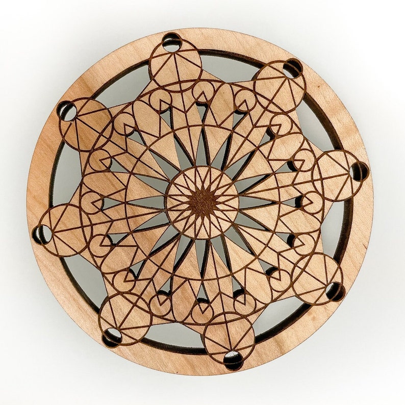 May include: A round, wooden coaster with a geometric design. The coaster features a central starburst pattern surrounded by interconnected circles and lines, all laser-cut into the light-colored wood. The design is intricate and symmetrical.
