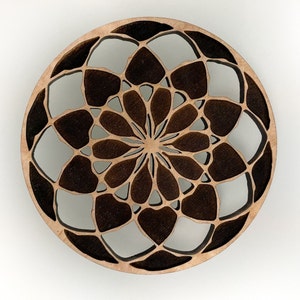 May include: A round, wooden coaster with a laser-cut floral design. The coaster features a light brown wood color with darker brown accents, creating a contrasting pattern. The design is symmetrical and intricate, with a central flower-like motif.