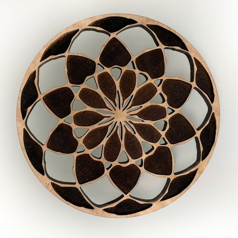 May include: A round, wooden coaster with a laser-cut floral design. The coaster features a light brown wood color with darker brown accents, creating a contrasting pattern. The design is symmetrical and intricate, with a central flower-like motif.