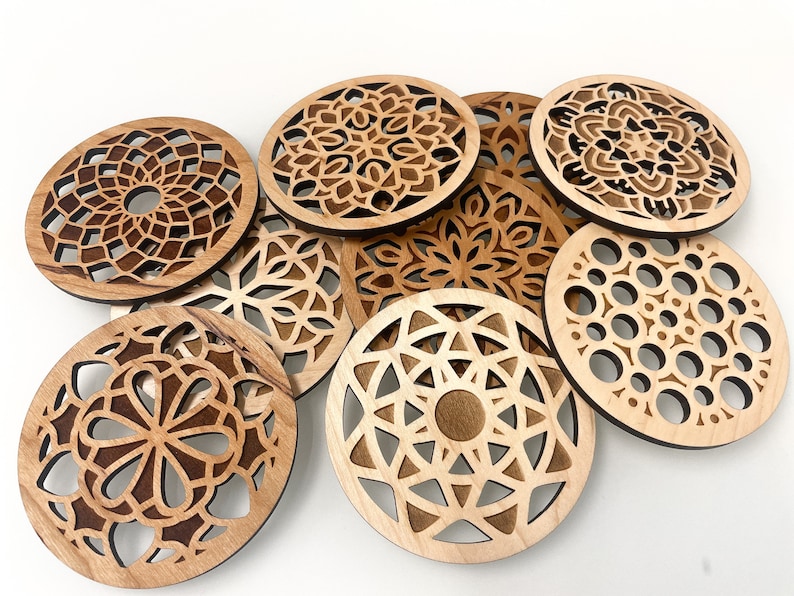 May include: Eight wooden coasters with intricate laser-cut mandala designs. The coasters are circular and feature various geometric patterns in shades of brown.