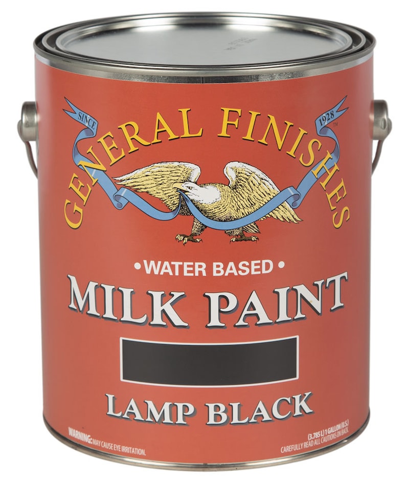 May include: A 1-gallon can of General Finishes Milk Paint in Lamp Black. The can is red with a white eagle and ribbon design. The text "General Finishes" is in a white script font. The text "Milk Paint" is in a black sans-serif font. The text "Lamp Black" is in a black sans-serif font. The text "Water Based" is in a black sans-serif font. The text "Since 1928" is in a white sans-serif font. The text "Warning: May Cause Eye Irritation" is in a black sans-serif font.