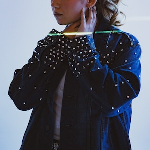 May include: A blue denim jacket with rhinestones on the sleeves and collar. The jacket is unbuttoned and the person wearing it is holding their hand to their neck.