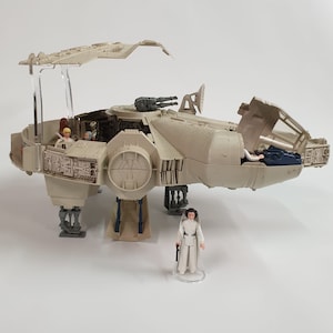 May include: A tan-colored toy Millennium Falcon spaceship with an open cockpit and various figures inside. A Princess Leia figure stands in front of the ship. The ship has a detailed design with visible guns and landing gear, and is set against a white background.