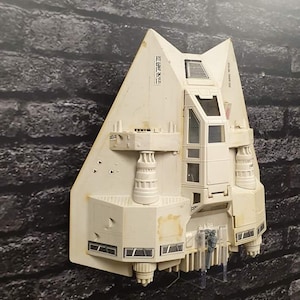 May include: Off-white model spacecraft with a triangular design and intricate detailing. The model shows signs of wear and tear. It is mounted on a dark brick wall. The model's dimensions are in centimeters.