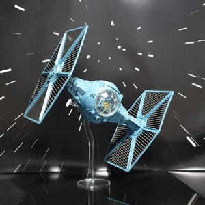 May include: A blue TIE fighter model with a clear display stand. The model is posed in flight with the wings extended. The background is a black and white pattern.