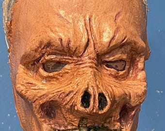 May include: A detailed, realistic-looking, orange-toned, latex Halloween mask with a skull-like face and a toothy grin. The mask has a textured surface and appears to be made of high-quality materials.