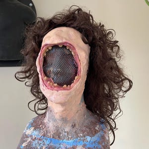 K&ouml;nnte beinhalten: Ein realistischer, blasser menschlicher Kopf mit langen, braunen, lockigen Haaren. Der Kopf hat einen gro&szlig;en, offenen Mund mit einem schwarzen Netz, das das Innere des Mundes bedeckt.