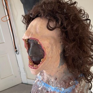 K&ouml;nnte beinhalten: Nahaufnahme einer realistischen Horror-Maske mit weit offenem Mund, die ein schwarzes Netz enth&uuml;llt, das das Innere des Mundes bedeckt. Die Maske hat einen blassen Hautton und ist aus Latex. Die Maske wird auf einem Mannequin mit braunen Locken getragen.
